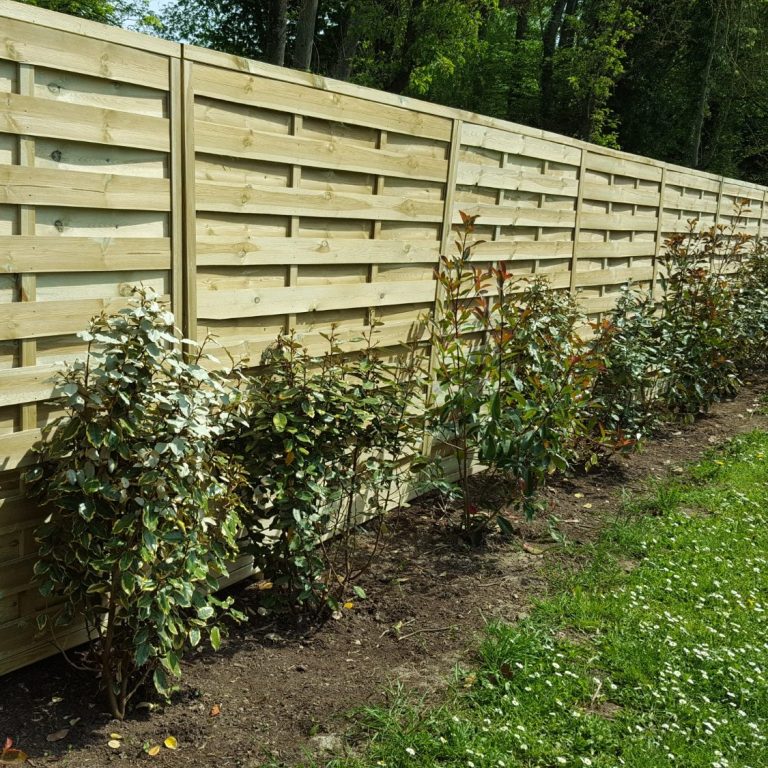 Clôture en bois avec des plantes le long de la base, dans un environnement verdoyant.